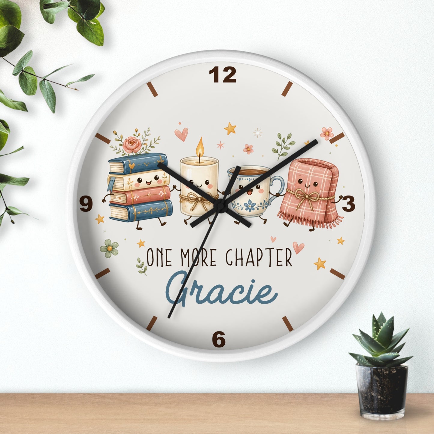 A round wall clock with a white frame and a whimsical book-themed face. The clock face features cute illustrations of a stack of books, a candle, a teacup, and a pink blanket, along with the text 'ONE MORE CHAPTER Gracie'.