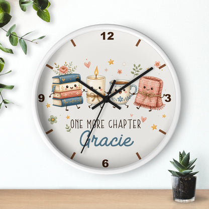 A round wall clock with a white frame and a whimsical book-themed face. The clock face features cute illustrations of a stack of books, a candle, a teacup, and a pink blanket, along with the text 'ONE MORE CHAPTER Gracie'.