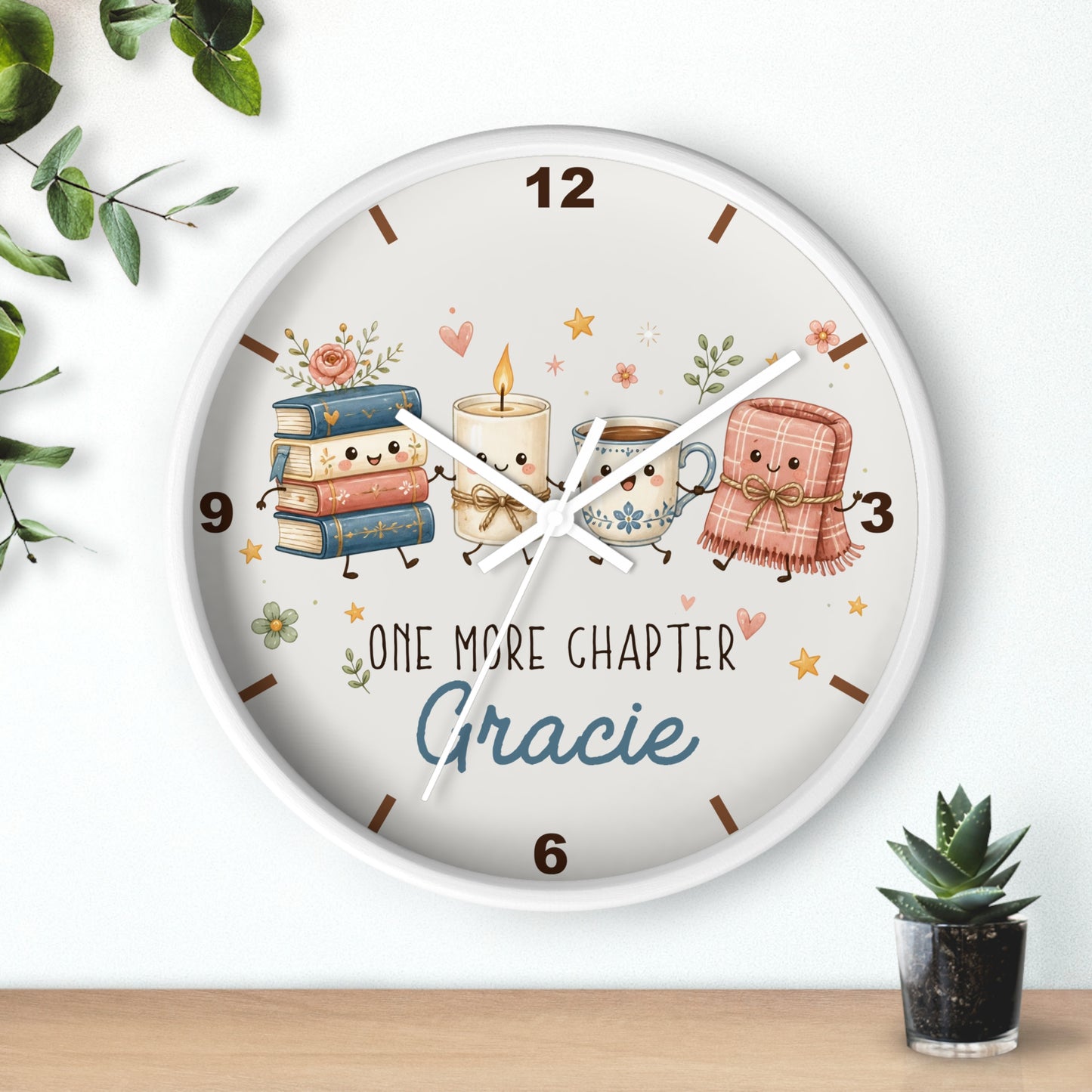 A round wall clock with a white frame and a whimsical illustration on the face showing a stack of books, a candle, a mug, and a pink blanket with the text 'ONE MORE CHAPTER Gracie'. The clock is mounted on a white wall above a light wood surface, with small green plants in the corners.