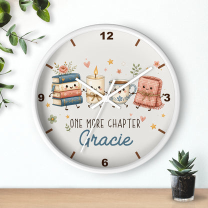 A round wall clock with a white frame and a whimsical illustration on the face showing a stack of books, a candle, a mug, and a pink blanket with the text 'ONE MORE CHAPTER Gracie'. The clock is mounted on a white wall above a light wood surface, with small green plants in the corners.