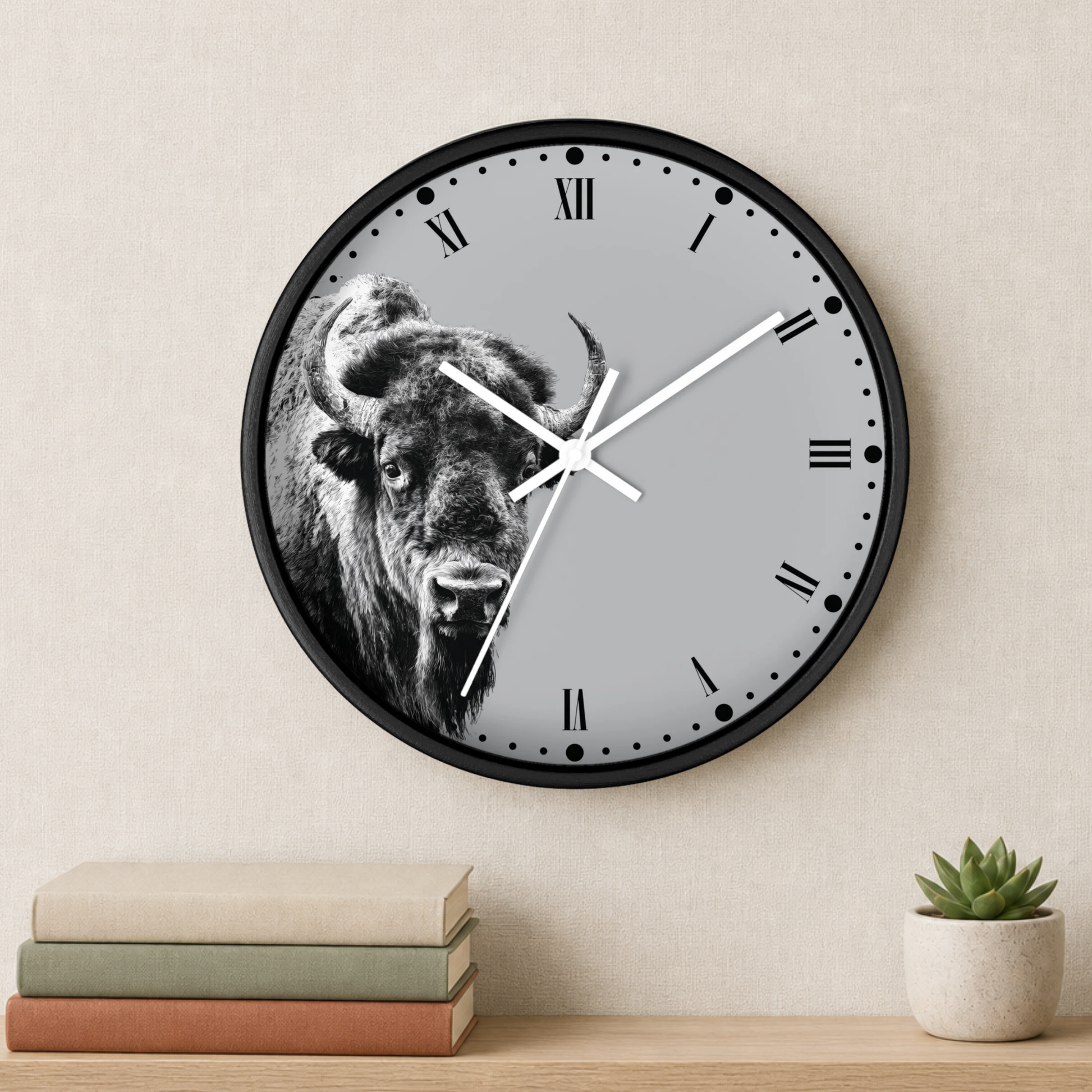 A round wall clock with a black frame and a gray face featuring a black-and-white bison illustration, mounted on a beige wall above a shelf with stacked books and a small potted succulent.
