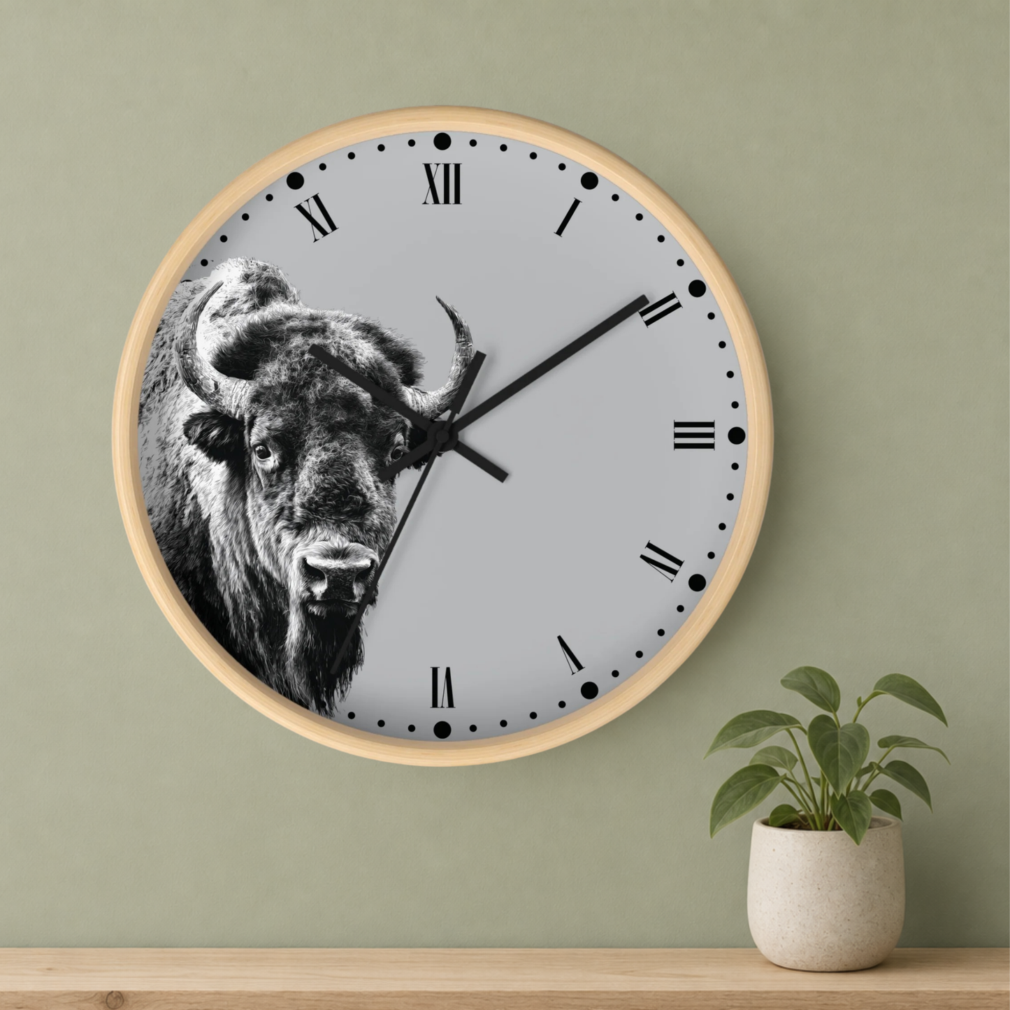 A round wall clock with a light wood frame and a black-and-white bison illustration on the clock face, mounted on a sage green wall above a wooden shelf with a small potted plant.