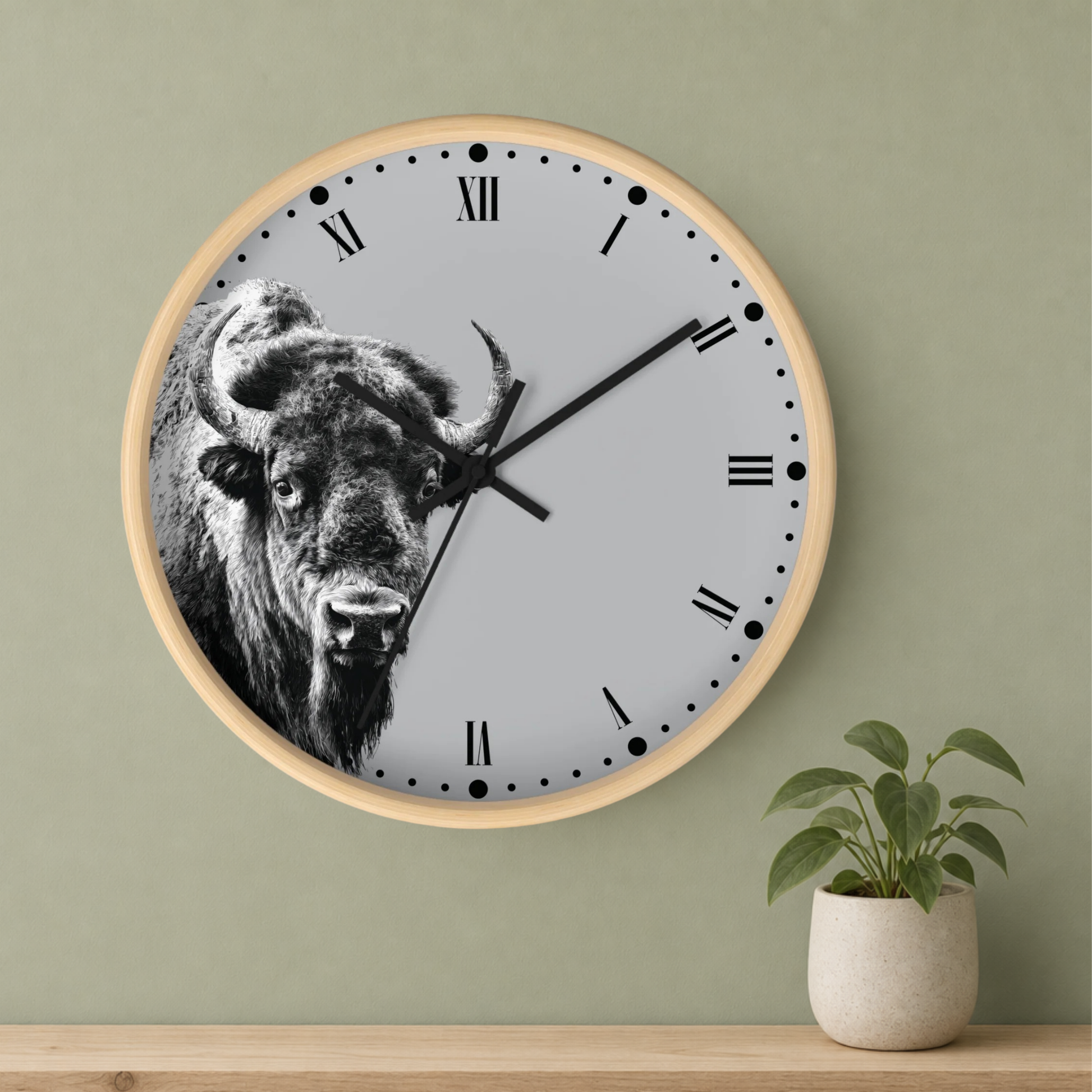 A round wall clock with a light wood frame and a black-and-white bison illustration on the clock face, mounted on a sage green wall above a wooden shelf with a small potted plant.