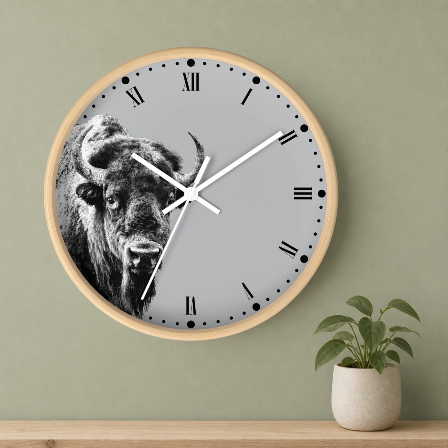 A round wall clock with a light wood frame and a gray face featuring a black-and-white bison illustration, mounted on a sage green wall above a wooden shelf with a small potted plant.