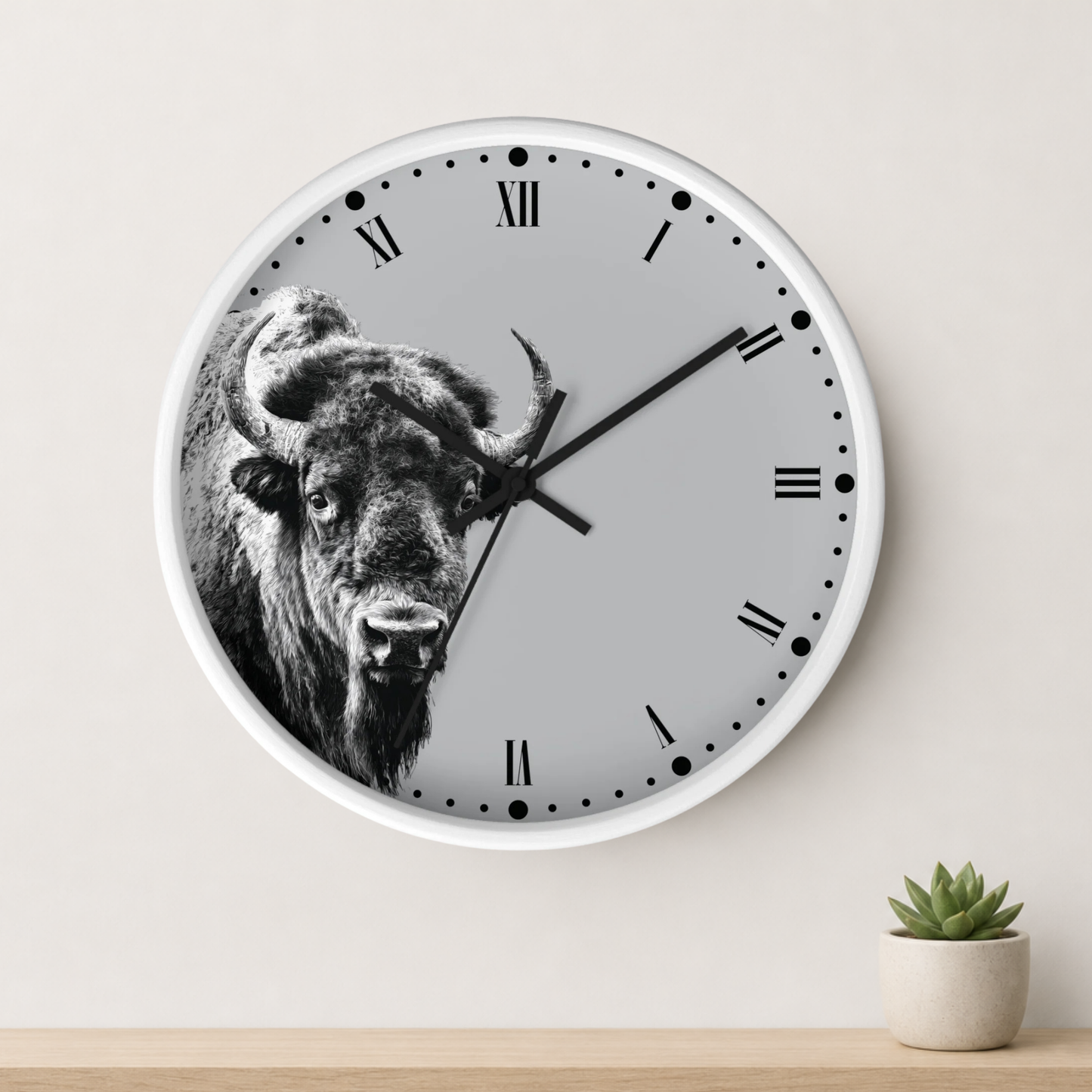 A round wall clock with a white frame and a black-and-white bison graphic on the face, mounted on a light beige wall above a wooden surface with a small potted succulent.