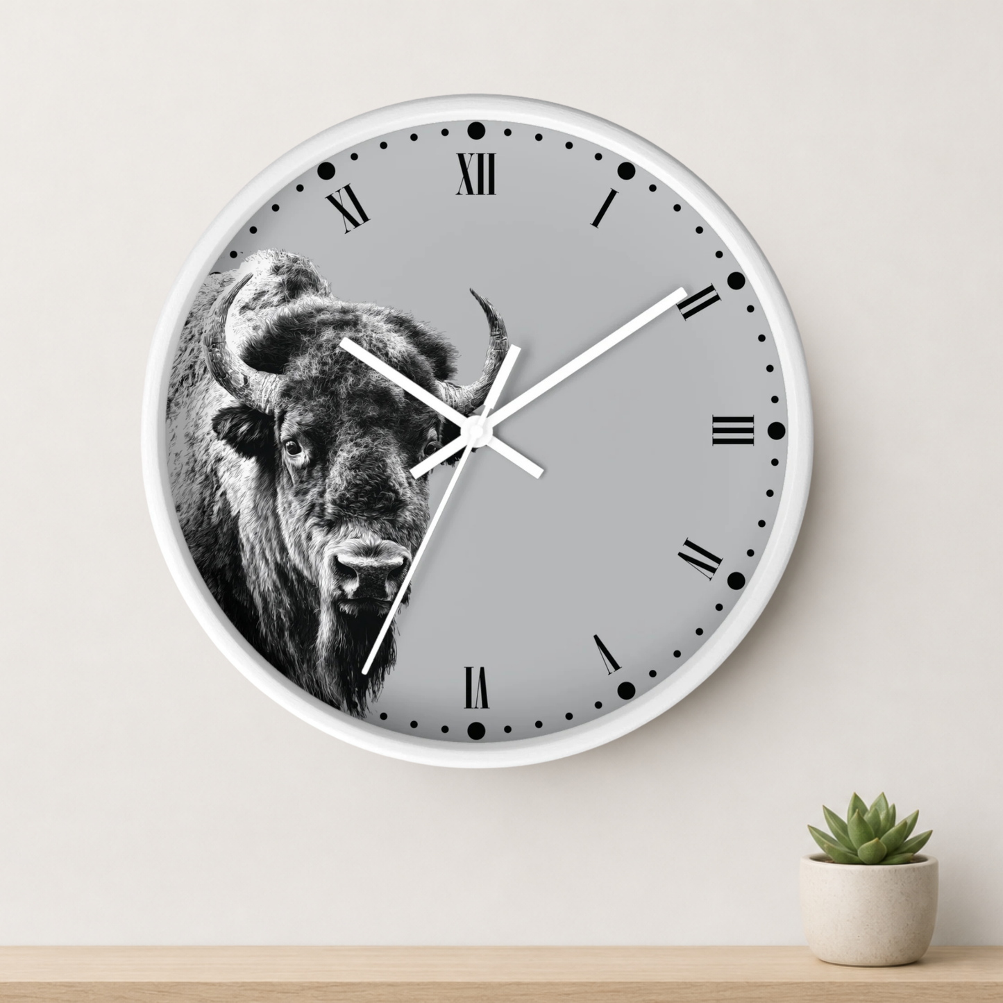 A round wall clock with a white frame and a gray face featuring a black-and-white bison illustration. The clock is mounted on a light beige wall above a wooden surface with a small potted succulent.