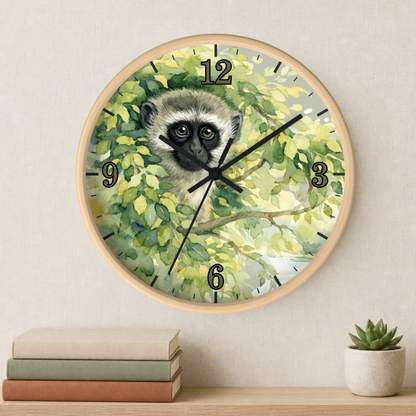 A round wall clock with a light wood frame and a printed face featuring a vervet monkey among green leaves, mounted on a beige wall above a shelf with stacked books and a small succulent.