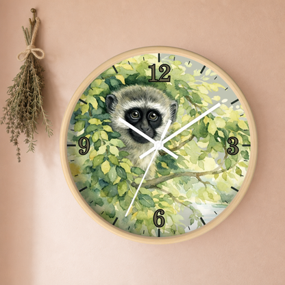 Round wall clock with a watercolor-style primate face among green leaves, mounted on a warm beige wall. The clock has a light wood-toned frame, black Arabic numerals, and white hands.