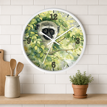 A round wall clock with a white frame hangs on a white subway-tile wall above a wooden shelf. The clock face features a painted vervet monkey surrounded by green leaves, with black Arabic numerals and white clock hands.