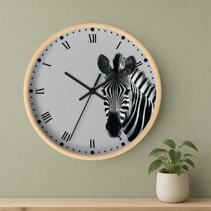 A round wall clock with a light wood frame and a white face featuring a black-and-white zebra illustration. The clock is mounted on a sage green wall above a wooden shelf with a small potted plant.