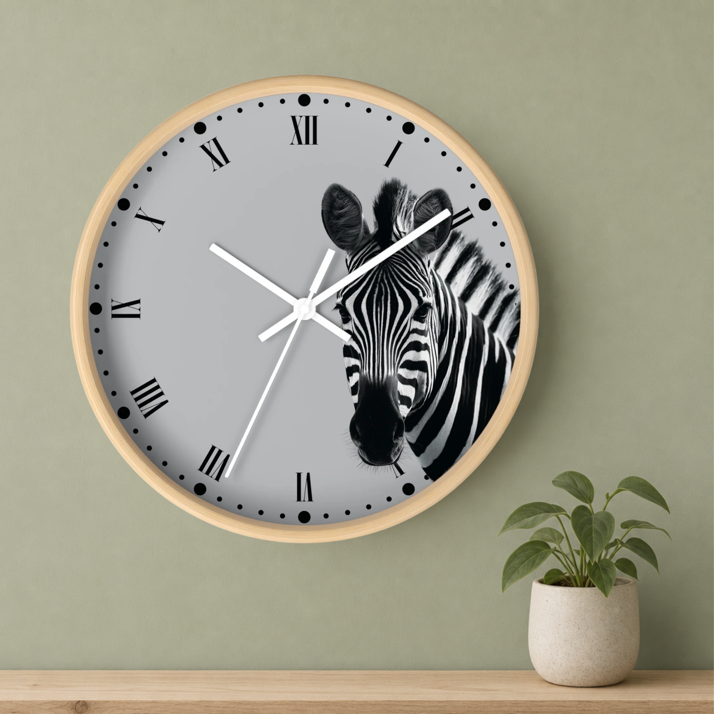 A round wall clock with a light wood frame and a gray face featuring a black-and-white zebra illustration, mounted on a sage green wall above a wooden shelf with a small potted plant.