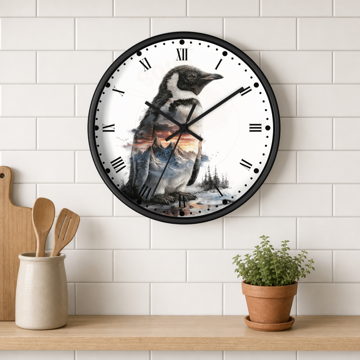 A round wall clock with a black frame and a penguin illustration over a mountain landscape on the clock face, mounted on a white subway tile wall above a wooden shelf with kitchen utensils and a potted plant.