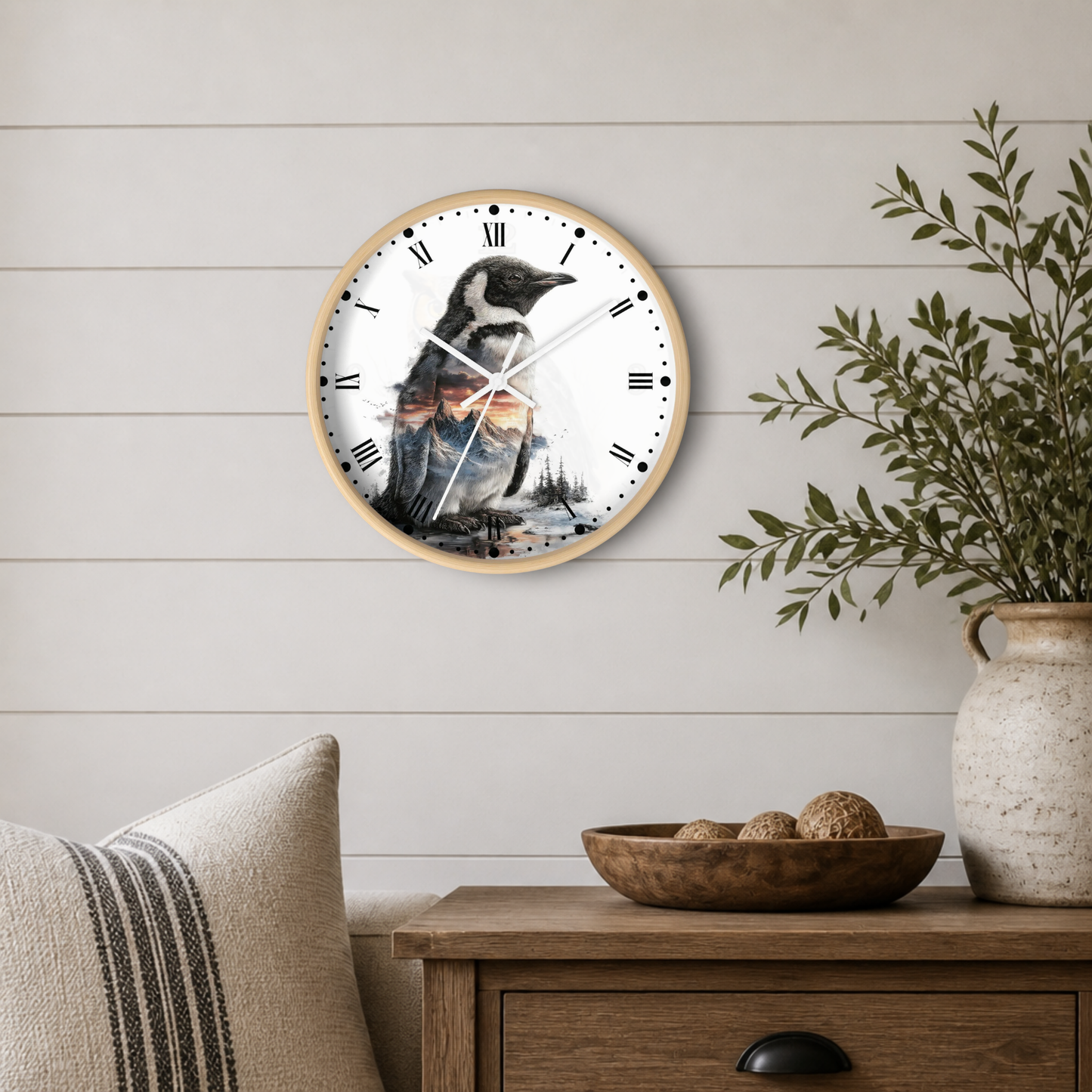 A round wall clock with a light wood frame hangs on a white shiplap wall above a wooden side table. The clock face features a detailed illustration of a penguin in black, white, and gray tones with a warm orange sunset in the background.