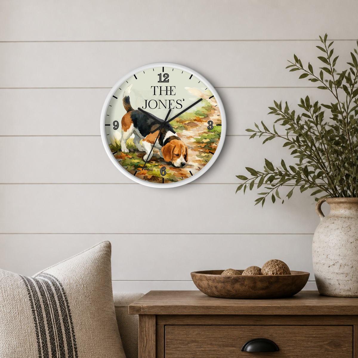 A personalized round wall clock featuring a beagle illustration and the text 'THE JONES', styled on a light wood paneled wall above a wooden side table with a vase and bowl.