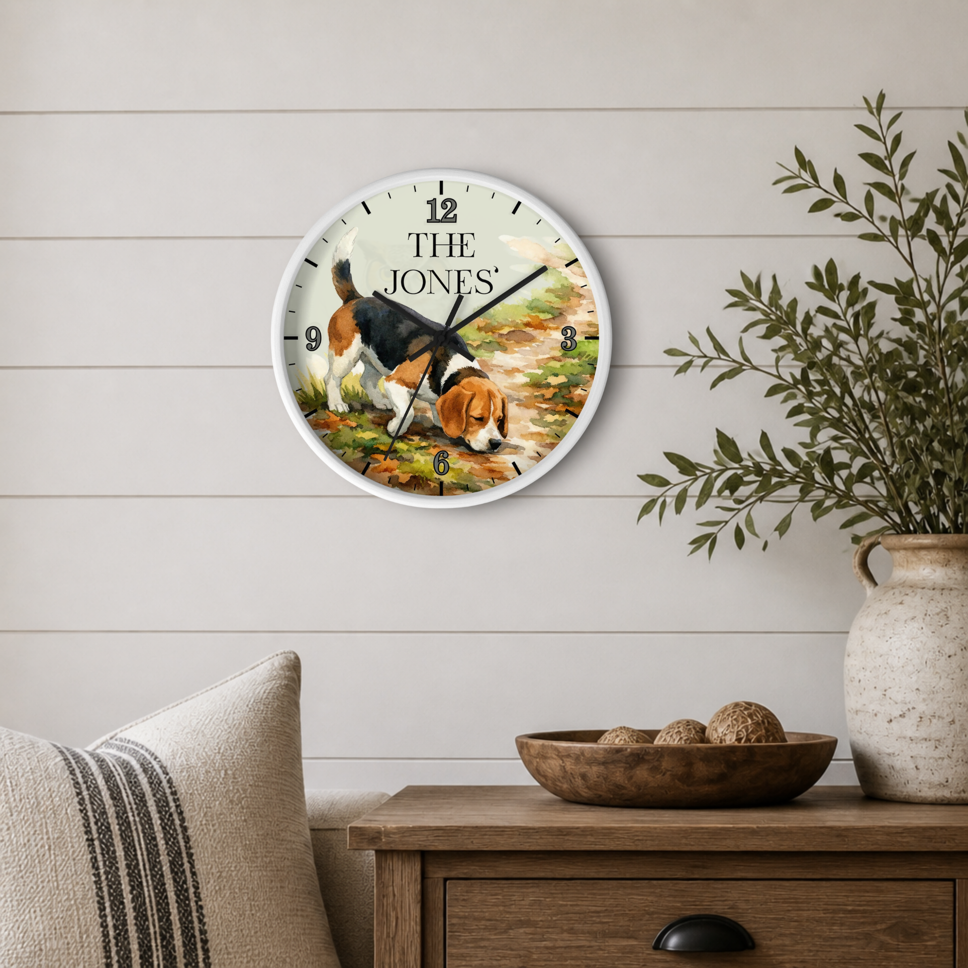 A personalized round wall clock featuring a beagle illustration and the text 'THE JONES', styled on a light wood paneled wall above a wooden side table with a vase and bowl.