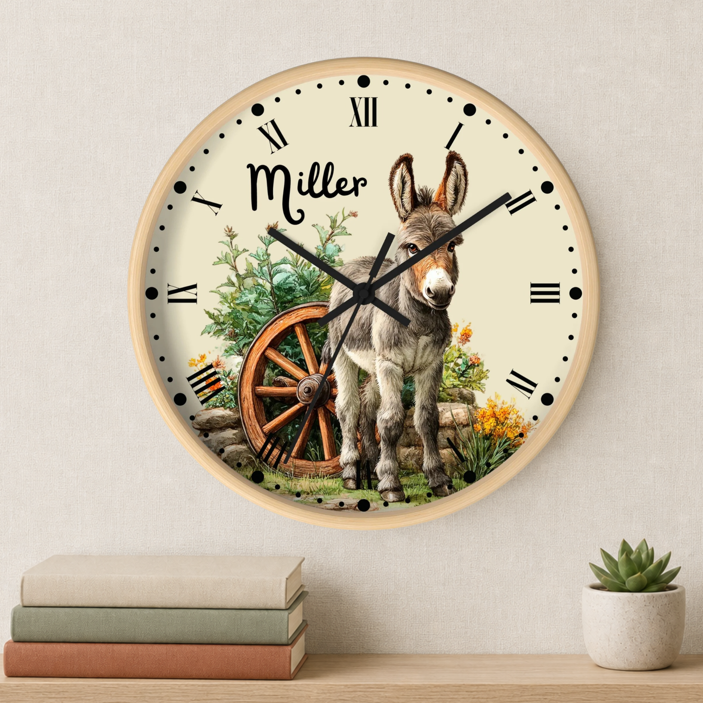 A personalized round wall clock with a light wood frame, featuring a cream background with black Roman numerals, black hands, and a rustic illustration of a donkey, a wagon wheel, and the name 'Miller' in script. The clock is shown hanging on a textured beige wall above a wooden shelf with stacked books and a small potted succulent.