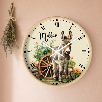 Personalized round wall clock featuring a donkey illustration, a rustic wagon wheel, and the name 'Miller' in script on a cream dial with black Roman numerals, mounted on a warm beige wall with dried herbs hanging to the left.