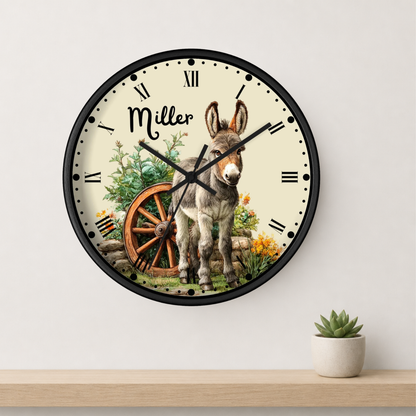 A round wall clock with a black frame and a cream-colored face featuring a vintage-style illustration of a donkey, a wooden wagon wheel, flowers, and the name 'Miller' in script. The clock is mounted on a light gray wall above a wooden shelf with a small potted succulent.