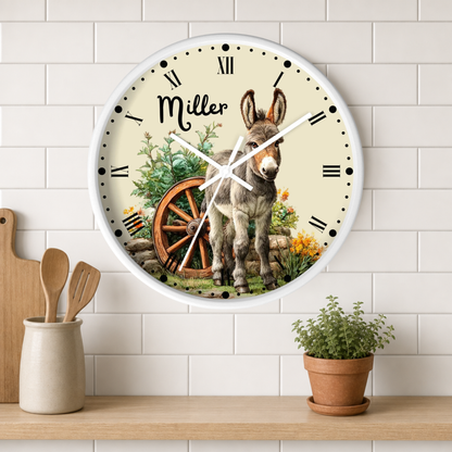 A personalized round wall clock with a white frame hangs on a white subway tile wall above a wooden shelf. The clock face features a cream background with black Roman numerals, white hands, and a detailed illustration of a donkey standing next to a wagon wheel, with the name 'Miller' written in script.