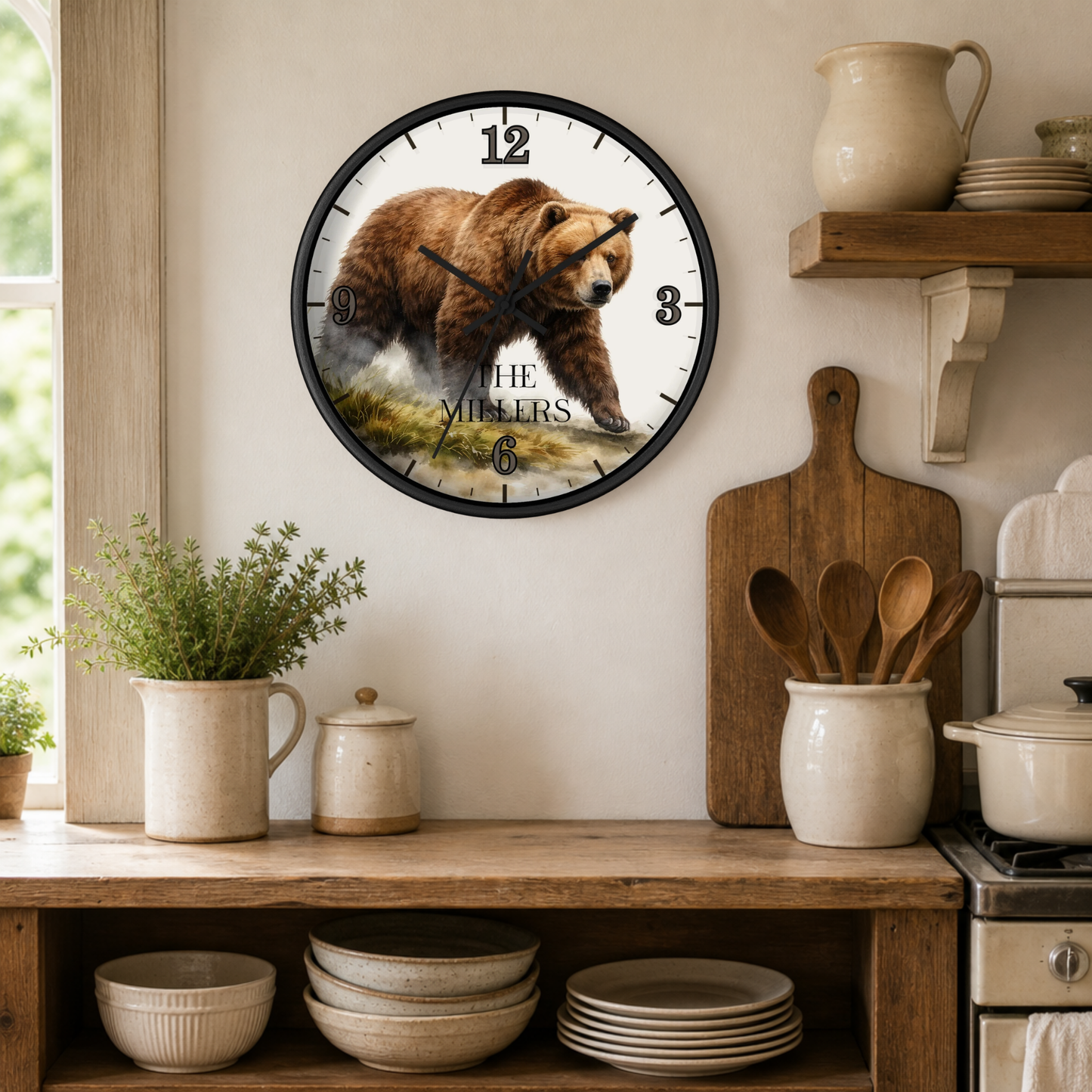 A round wall clock with a black frame hangs on a beige wall in a rustic kitchen setting. The clock face features a detailed illustration of a brown bear walking through grass, with the text 'THE MILLERS' printed below.