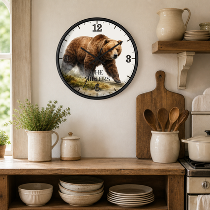 A round wall clock with a black frame hangs on a beige wall in a rustic kitchen setting. The clock face features a detailed illustration of a brown bear walking through grass, with the text 'THE MILLERS' printed below.