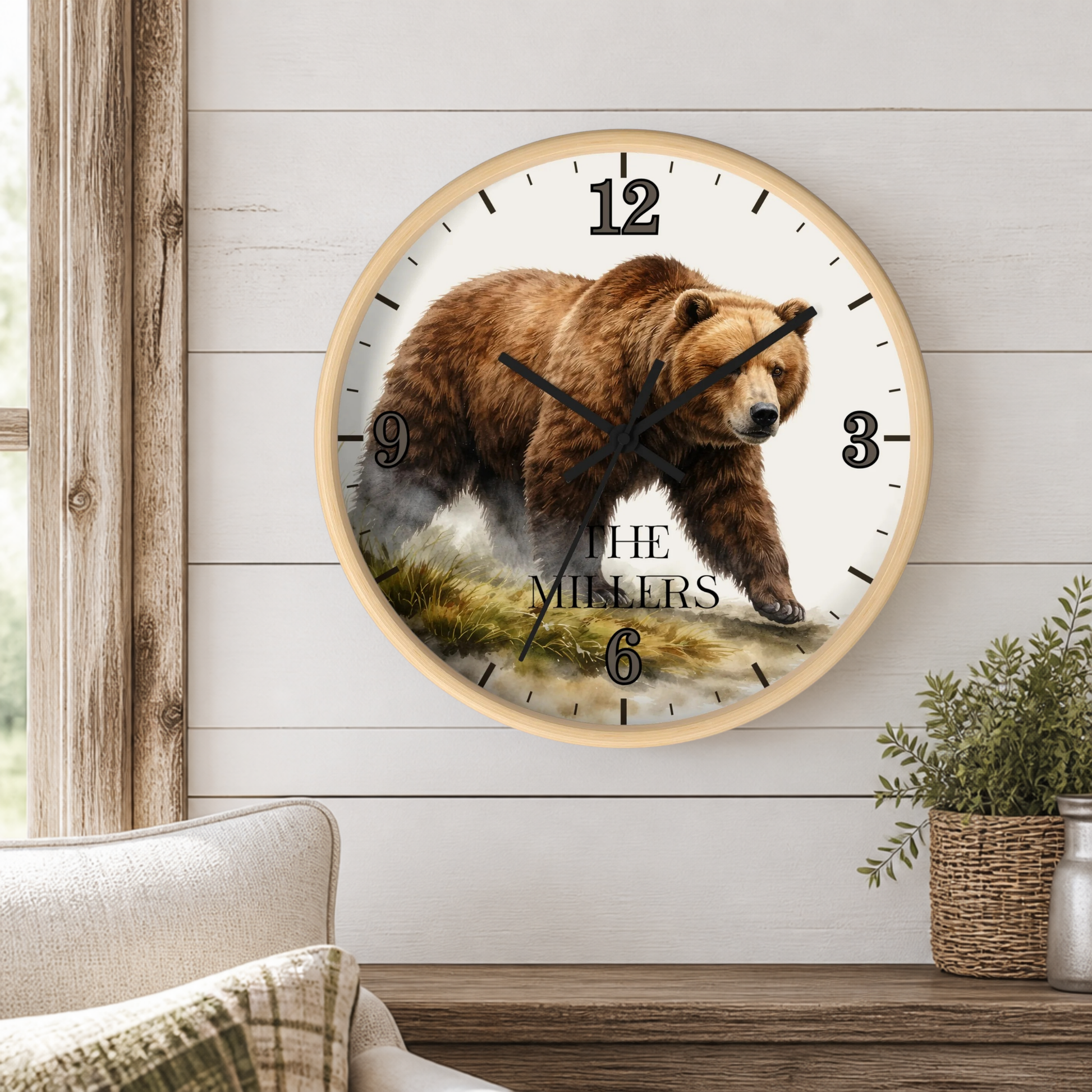 A round wall clock with a light wood frame hangs on a white shiplap wall above a wooden table. The clock face features a detailed brown bear illustration with the text 'THE MILLERS' and black Roman numerals.