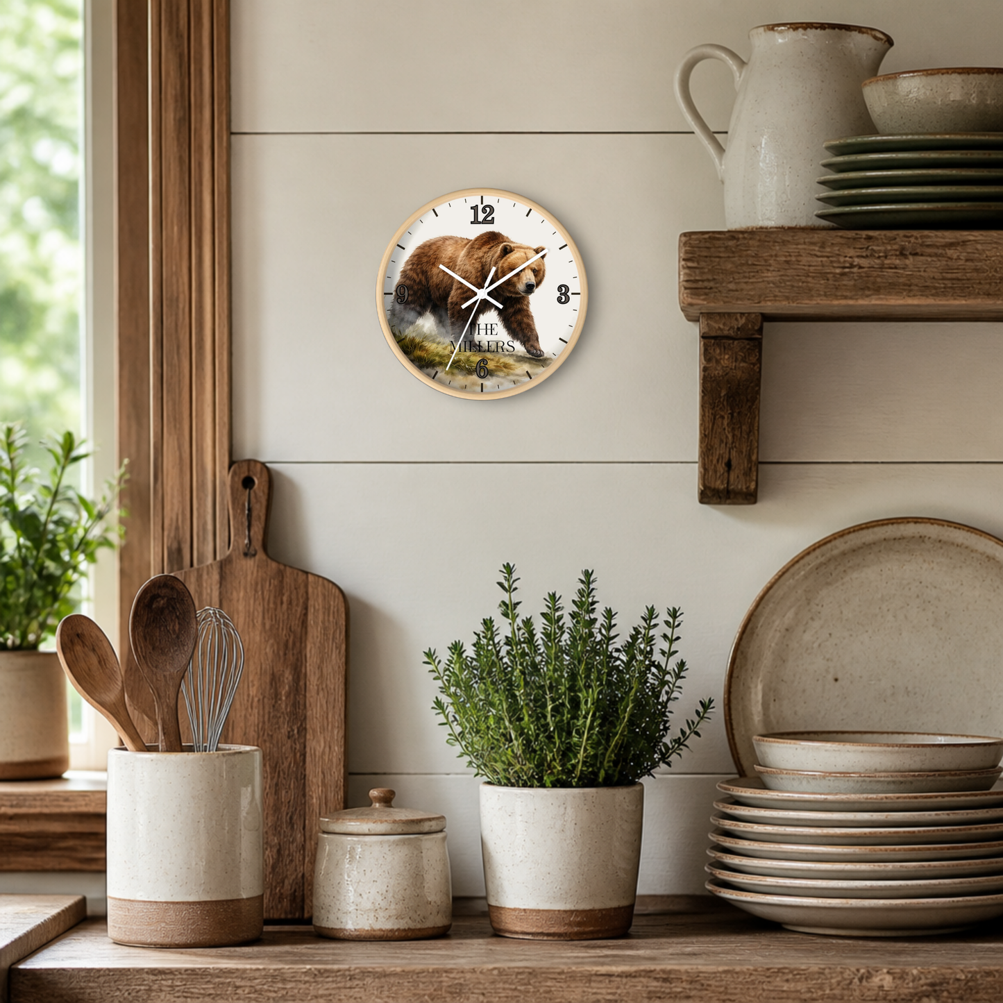A round wall clock featuring a brown bear illustration and the text 'The Bear & The Bee' on the wall in a rustic kitchen setting with wooden shelves, ceramic dishes, and potted plants.