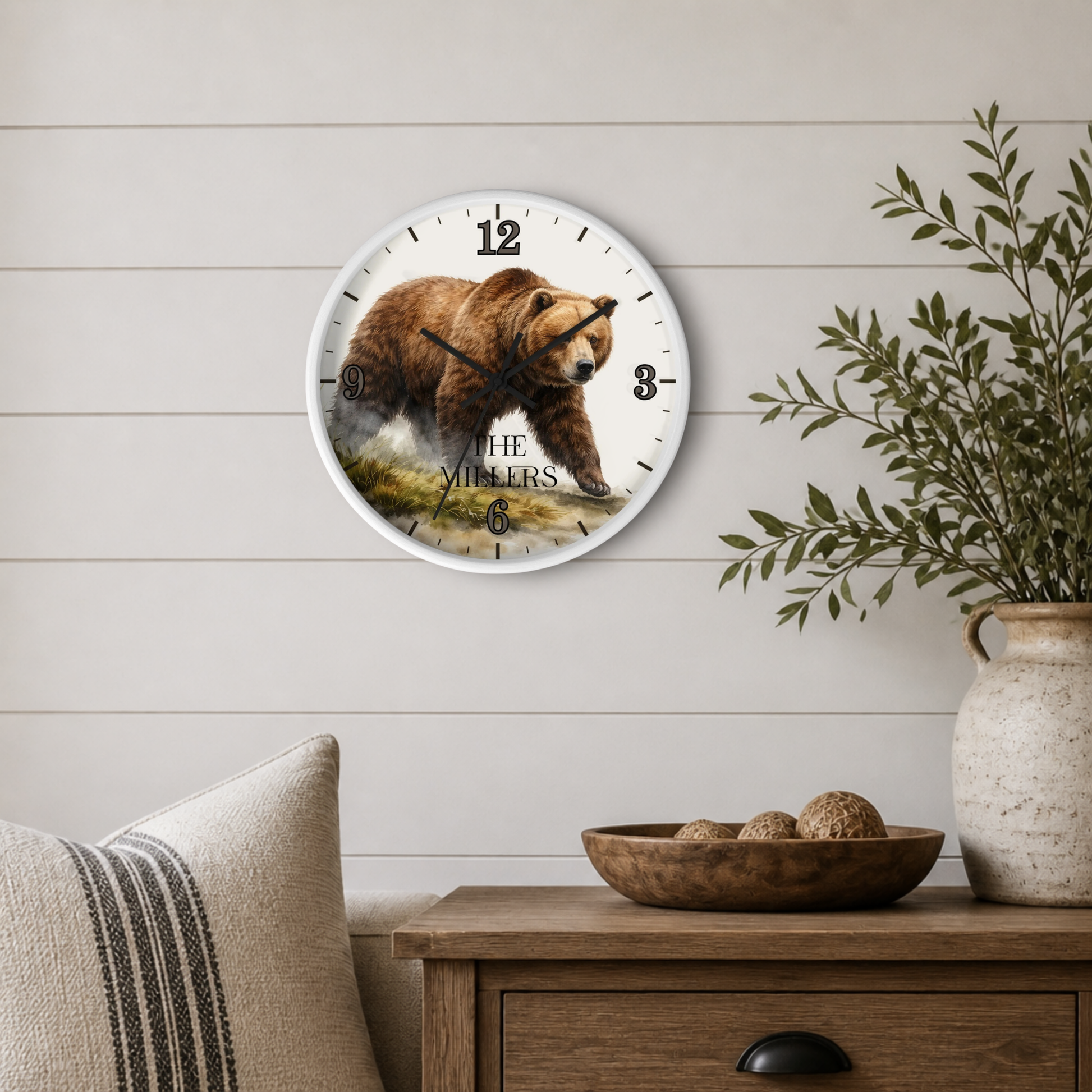 A round wall clock with a white frame hangs on a light wood shiplap wall above a wooden side table. The clock face features a detailed illustration of a brown grizzly bear walking through water, with the text 'THE MILLERS' printed below.
