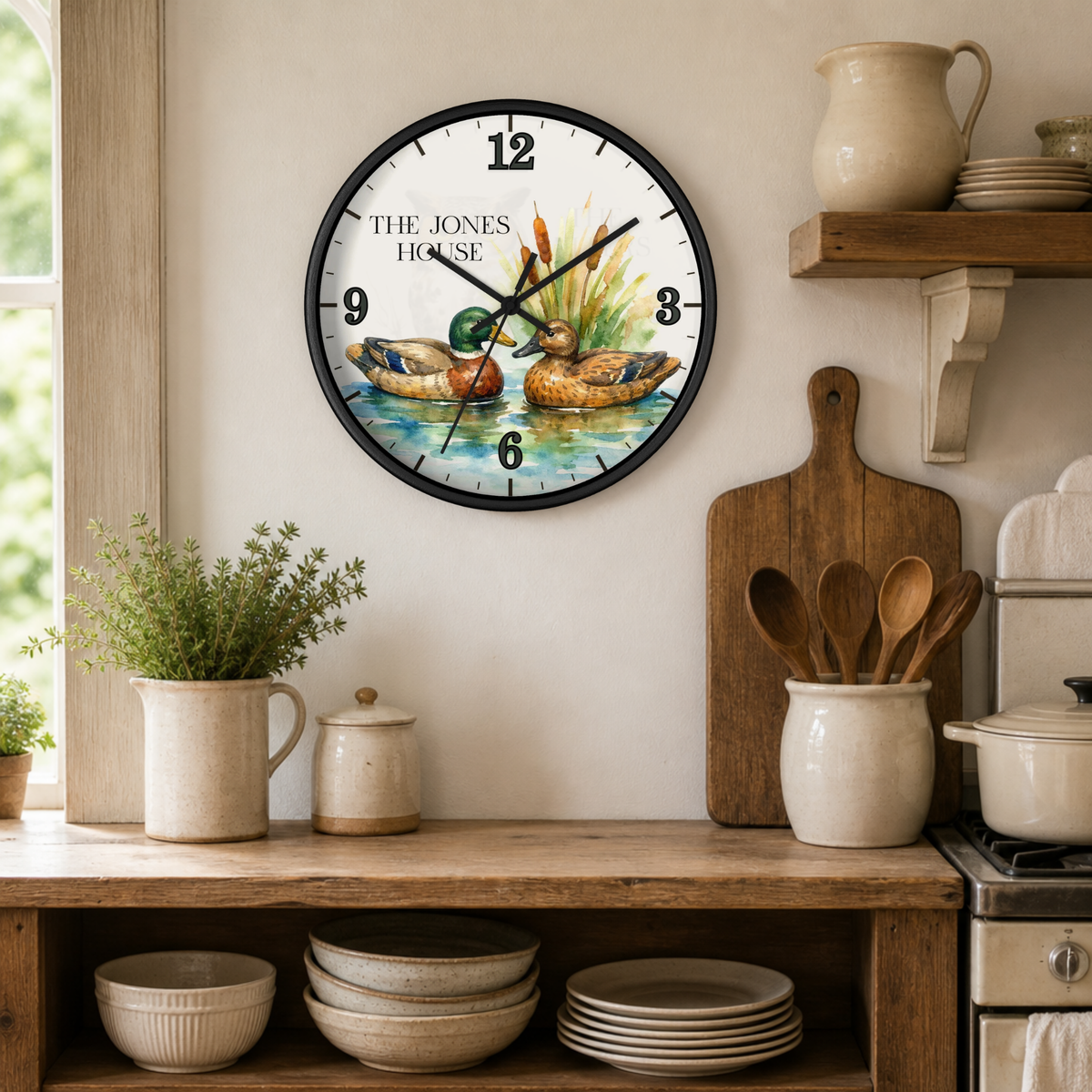 A round wall clock with a black frame hangs on a cream kitchen wall above a wooden counter. The clock face features a vintage-style illustration of two ducks in a pond with the text 'THE JONES HOUSE' at the top.