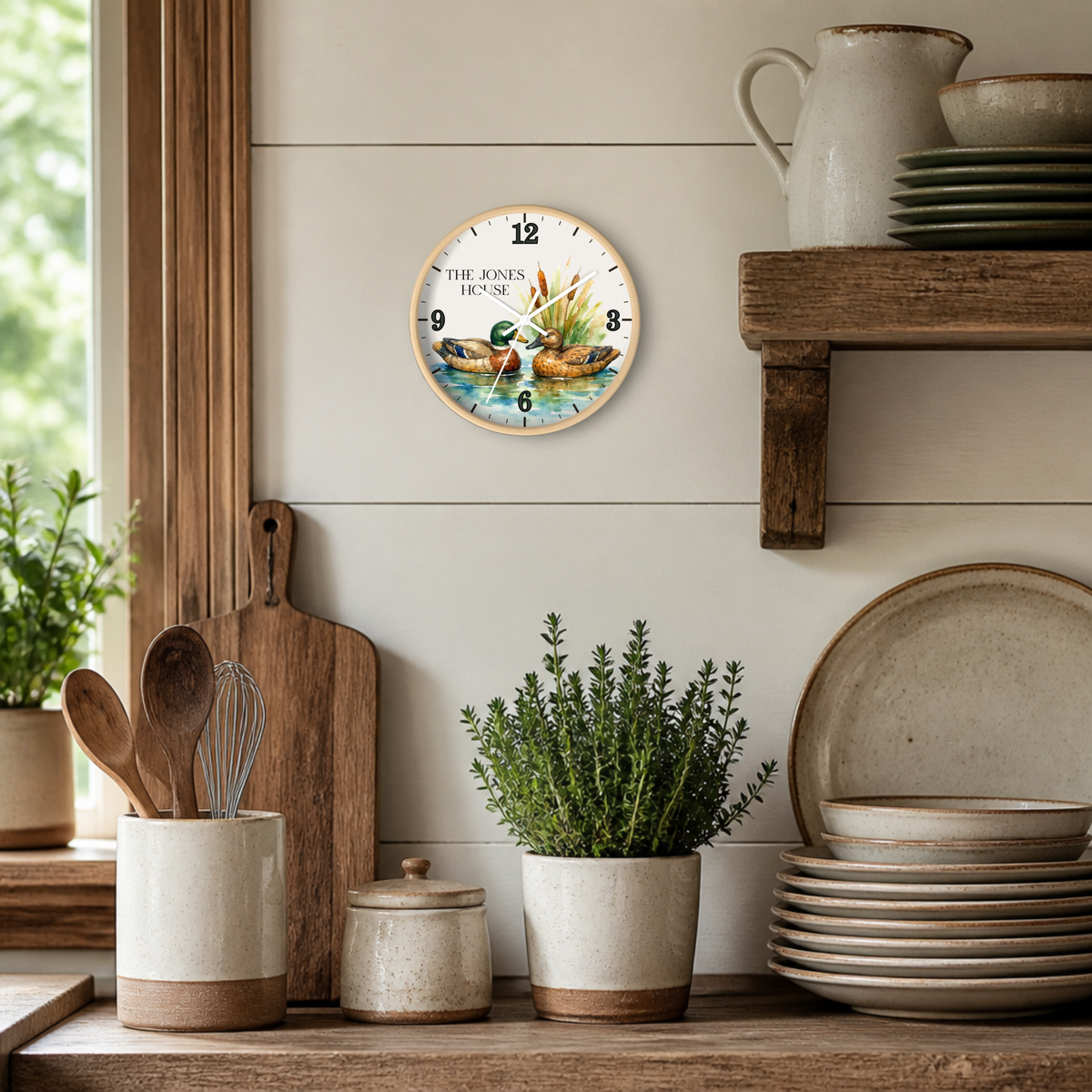 A round wall clock with a rustic duck illustration and the text 'THE JONES HOUSE' hangs on a white shiplap wall in a styled kitchen setting with wooden shelves, ceramic dishes, and potted plants.