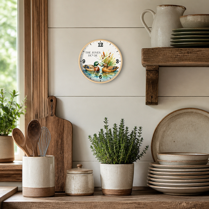 A round wall clock with a rustic duck illustration and the text 'THE JONES HOUSE' hangs on a white shiplap wall in a styled kitchen setting with wooden shelves, ceramic dishes, and potted plants.