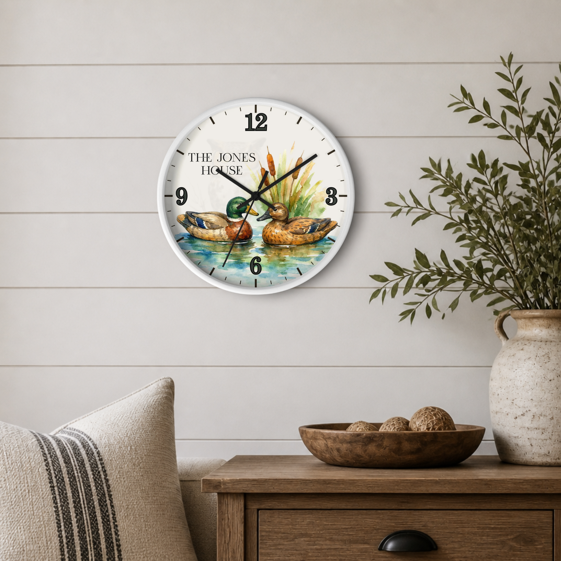 A round wall clock with a white face and black numerals, featuring an illustration of two ducks in a pond with the text 'THE JONES HOUSE' on the clock face. The clock is mounted on a light beige shiplap wall above a wooden side table with a vase of greenery and a pillow.
