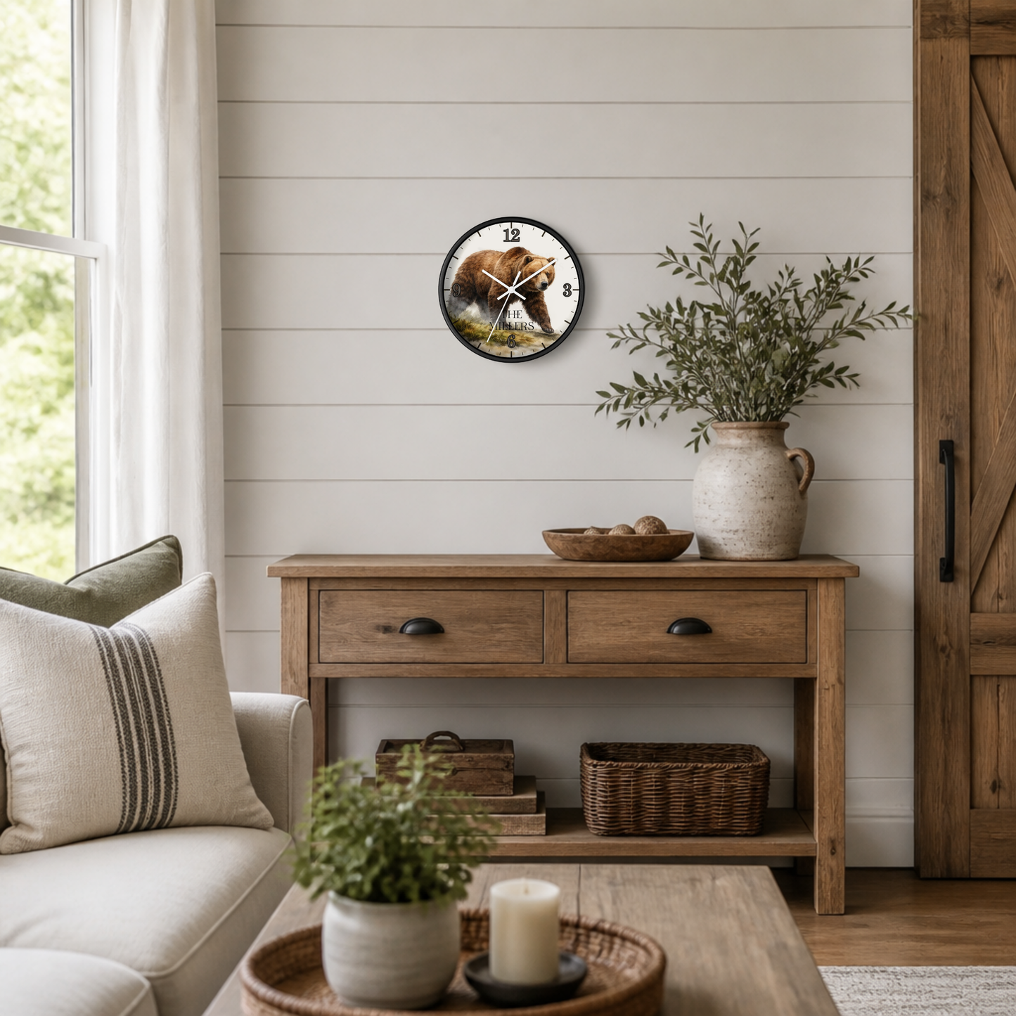 A round wall clock with a rustic illustration of a bear hanging on a white shiplap wall above a wooden console table in a styled living room.