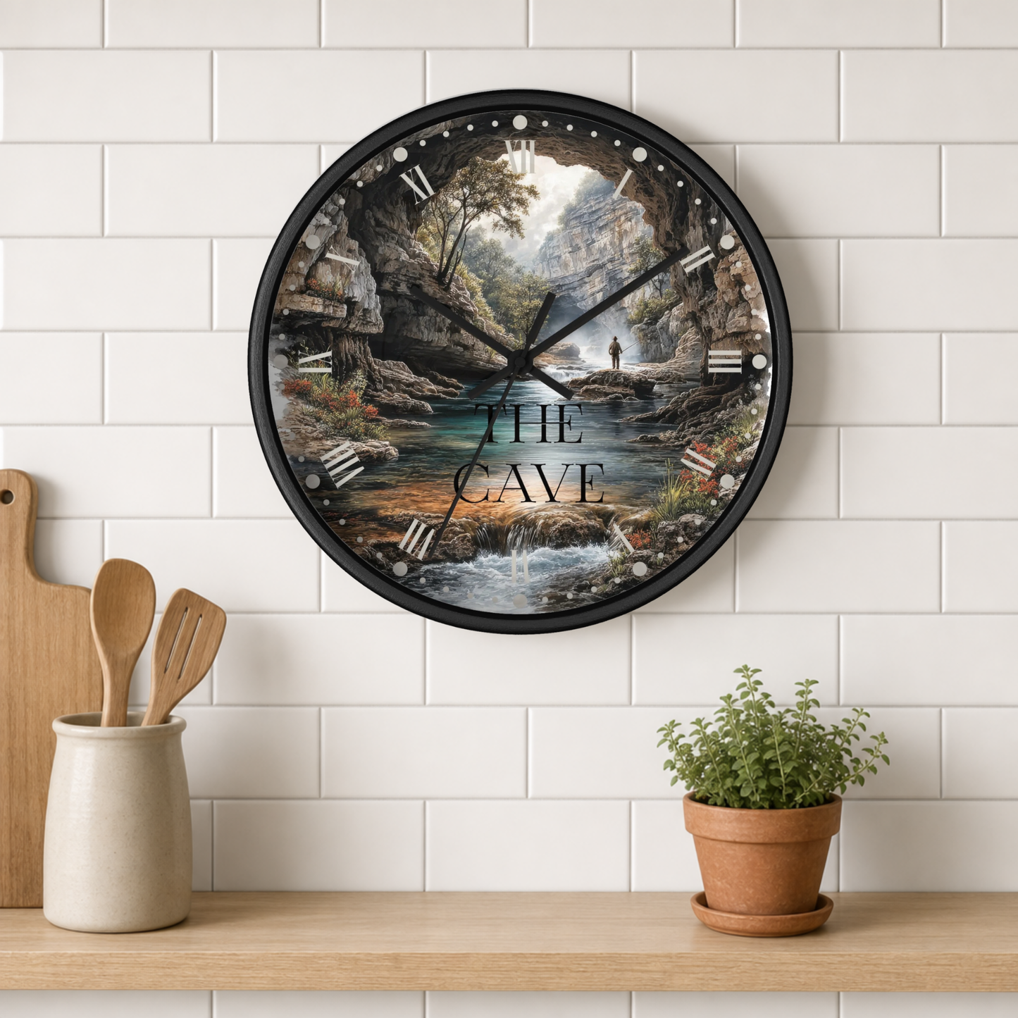 A round wall clock with a black frame and a detailed cave landscape print, mounted on a white subway tile wall above a wooden shelf with kitchen utensils and a potted plant.