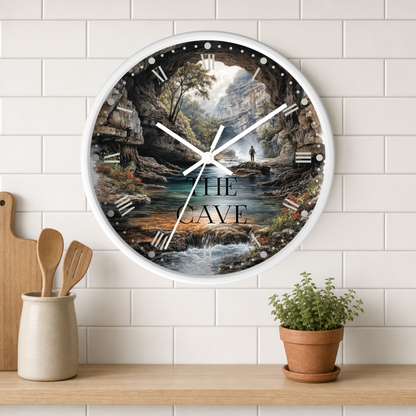 Round white wood wall clock with a detailed illustration of an angler in a stream fishing and the text 'The Cave' hanging on a white tiled kitchen wall above a counter with cooking utensils and a small potted plant.