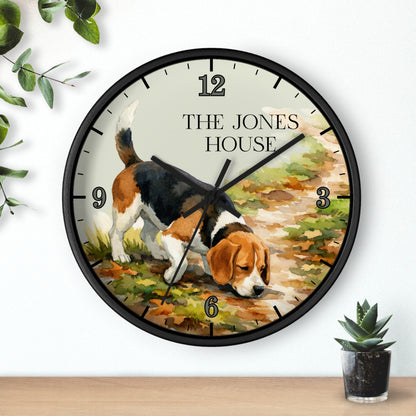 A round wall clock with a black frame and a printed illustration of a beagle dog sniffing the ground, with the text 'THE JONES HOUSE' at the top. The clock is mounted on a white wall above a wooden surface, with small green plants in the corners.