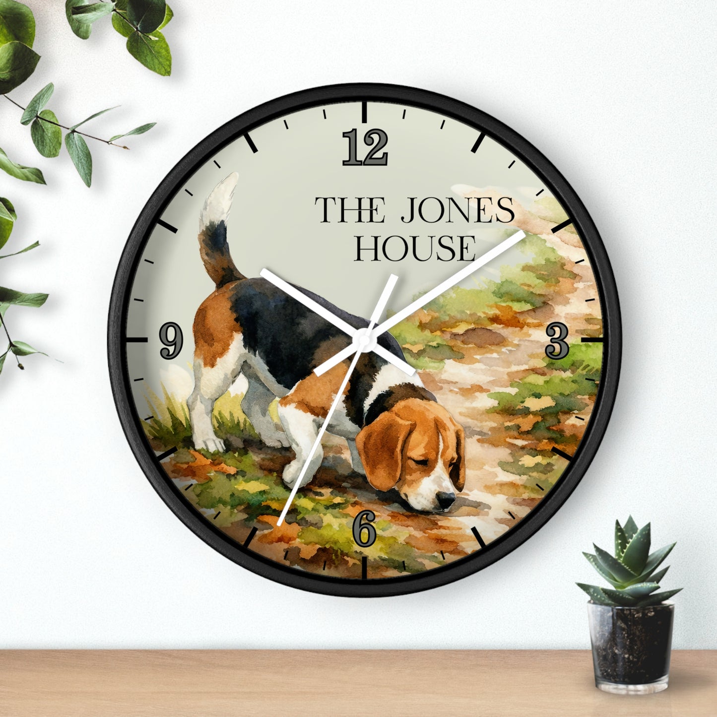 A round wall clock with a black frame and a printed illustration of a beagle dog on the face, with the text 'THE JONES HOUSE' at the top. The clock is shown hanging on a white wall above a wooden surface, with small green plants in the corners.