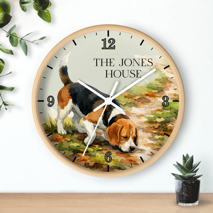 A round wall clock with a light wood frame, featuring a printed illustration of a beagle dog on a path and the text 'THE JONES HOUSE' on the clock face. The clock is shown hanging on a white wall above a wooden surface with small potted plants nearby.