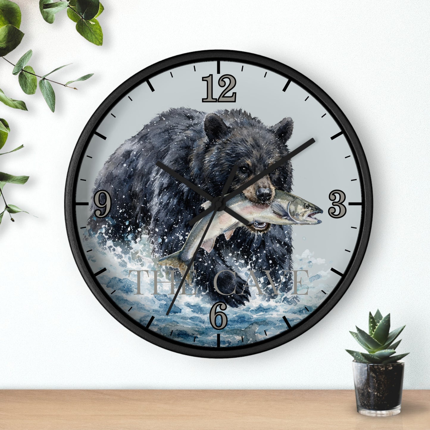 A round wall clock with a black frame and a printed design of a black bear carrying a fish across blue water, mounted on a white wall above a wooden surface with small green plants.