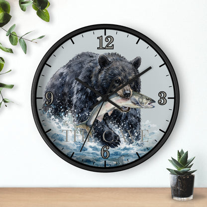 A round wall clock with a black frame and a printed design of a black bear carrying a fish across blue water, mounted on a white wall above a wooden surface with small green plants.