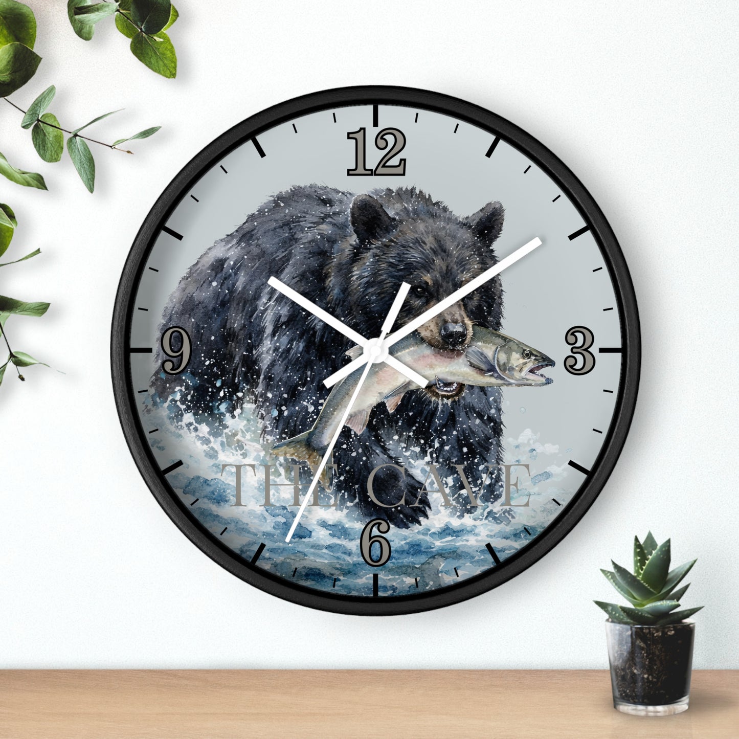 A round wall clock with a black frame and a printed illustration of a bear catching a fish, mounted on a white wall above a wooden surface with small green plants.