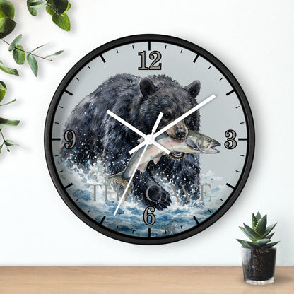 A round wall clock with a black frame and a printed illustration of a bear catching a fish, mounted on a white wall above a wooden surface with small green plants.