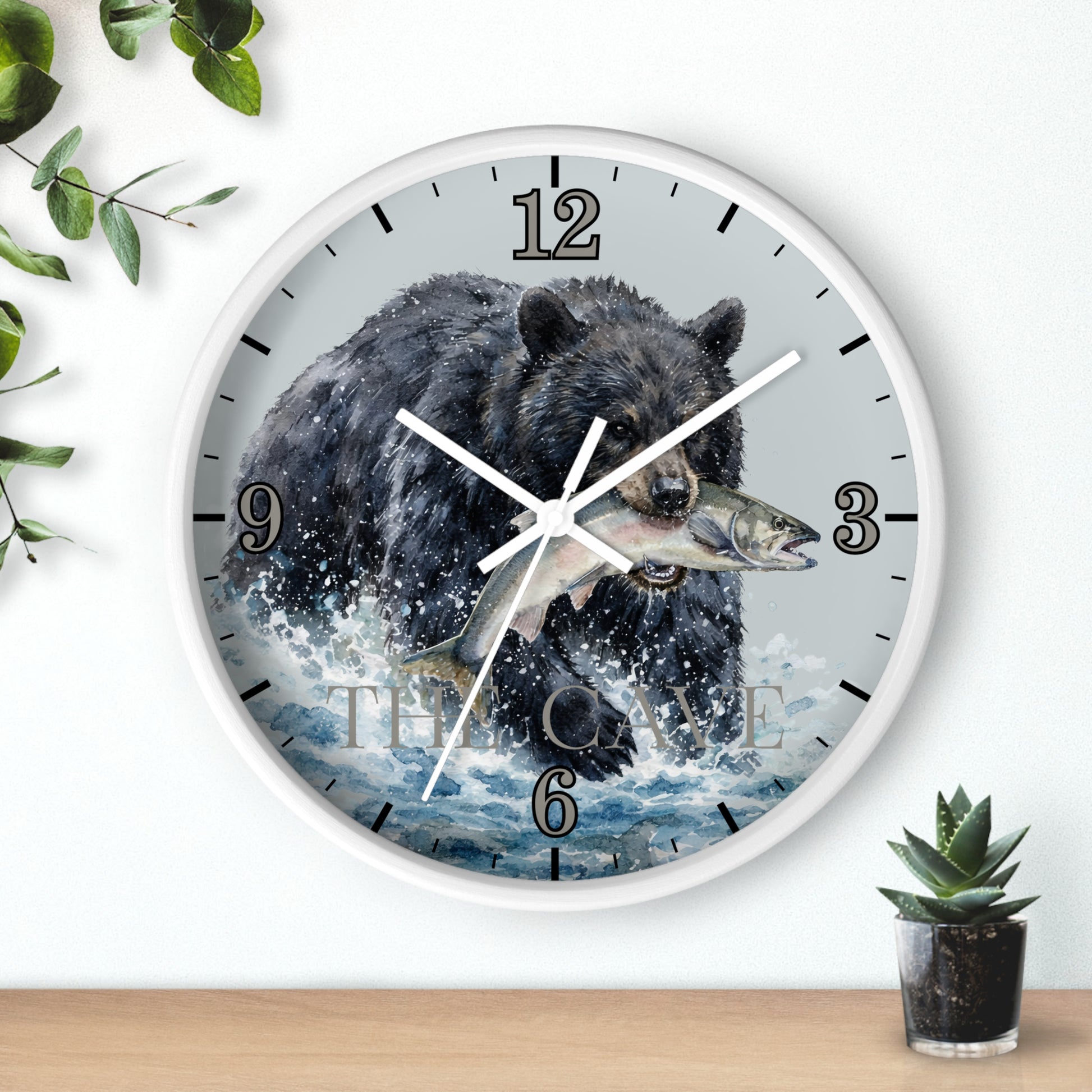 A round wall clock with a white frame and a printed design of a black bear catching a fish in splashing water. The clock is mounted on a white wall above a light wood surface, with small green plants in the corners.
