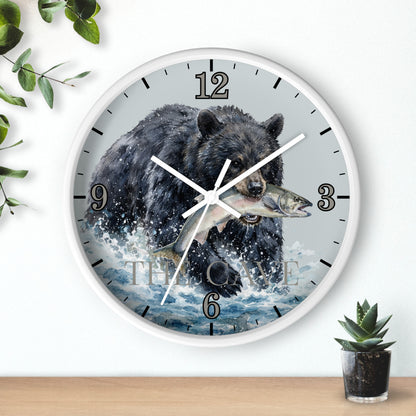 A round wall clock with a white frame and a printed design of a black bear catching a fish in splashing water. The clock is mounted on a white wall above a light wood surface, with small green plants in the corners.
