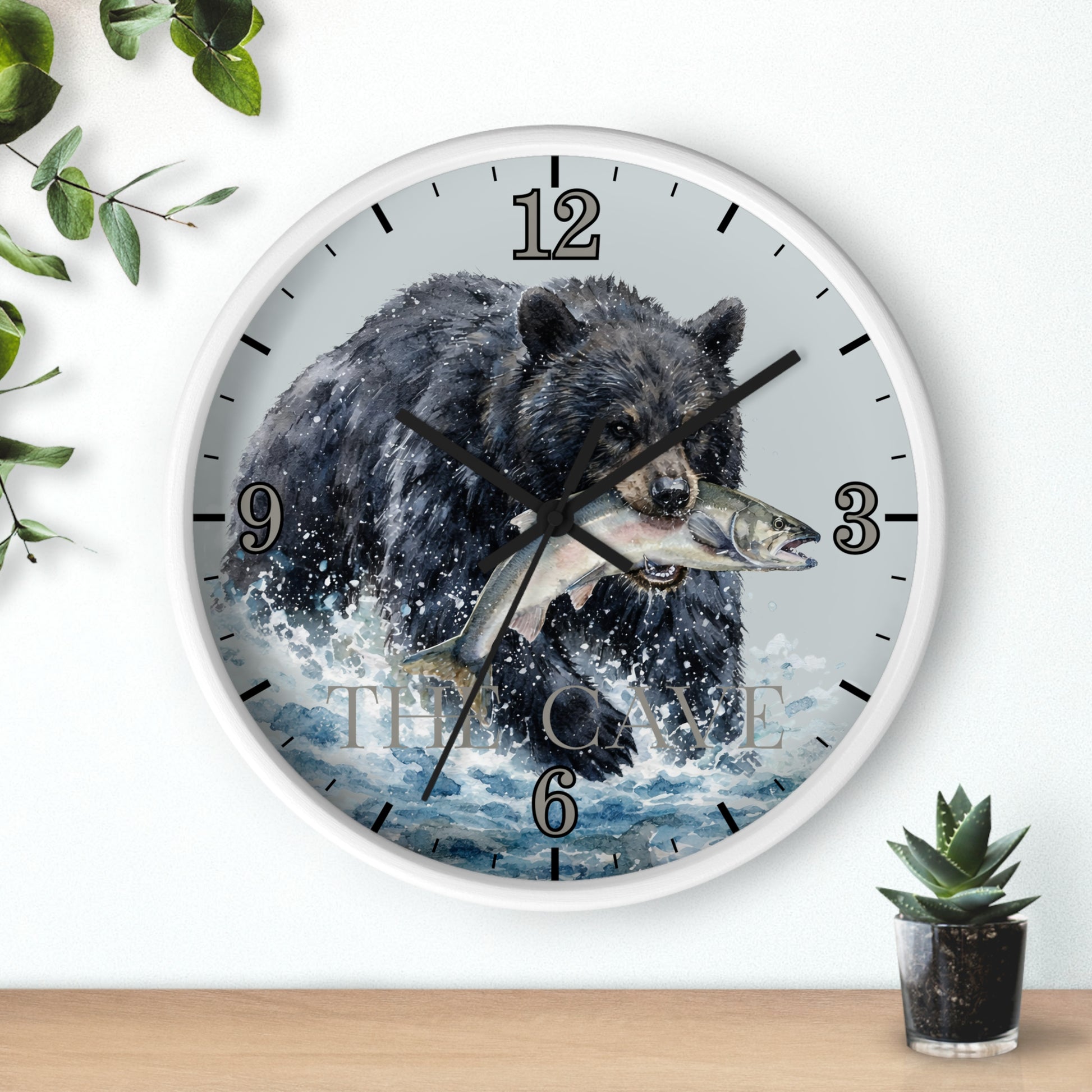 A round wall clock with a white frame and a printed design of a black bear catching a fish in splashing water. The clock is mounted on a white wall above a light wood surface, with decorative green leaves and a small potted succulent nearby.
