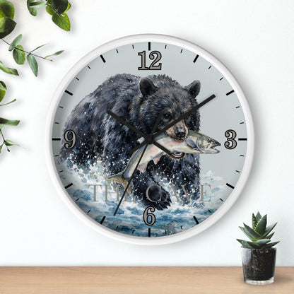 A round wall clock with a white frame and a printed design of a black bear catching a fish in splashing water. The clock is mounted on a white wall above a light wood surface, with decorative green leaves and a small potted succulent nearby.