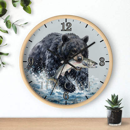 A round wall clock with a light wood frame, featuring a printed design of a black bear catching a fish in splashing water. The clock is mounted on a white wall above a wooden shelf, with decorative green leaves and a small potted succulent nearby.