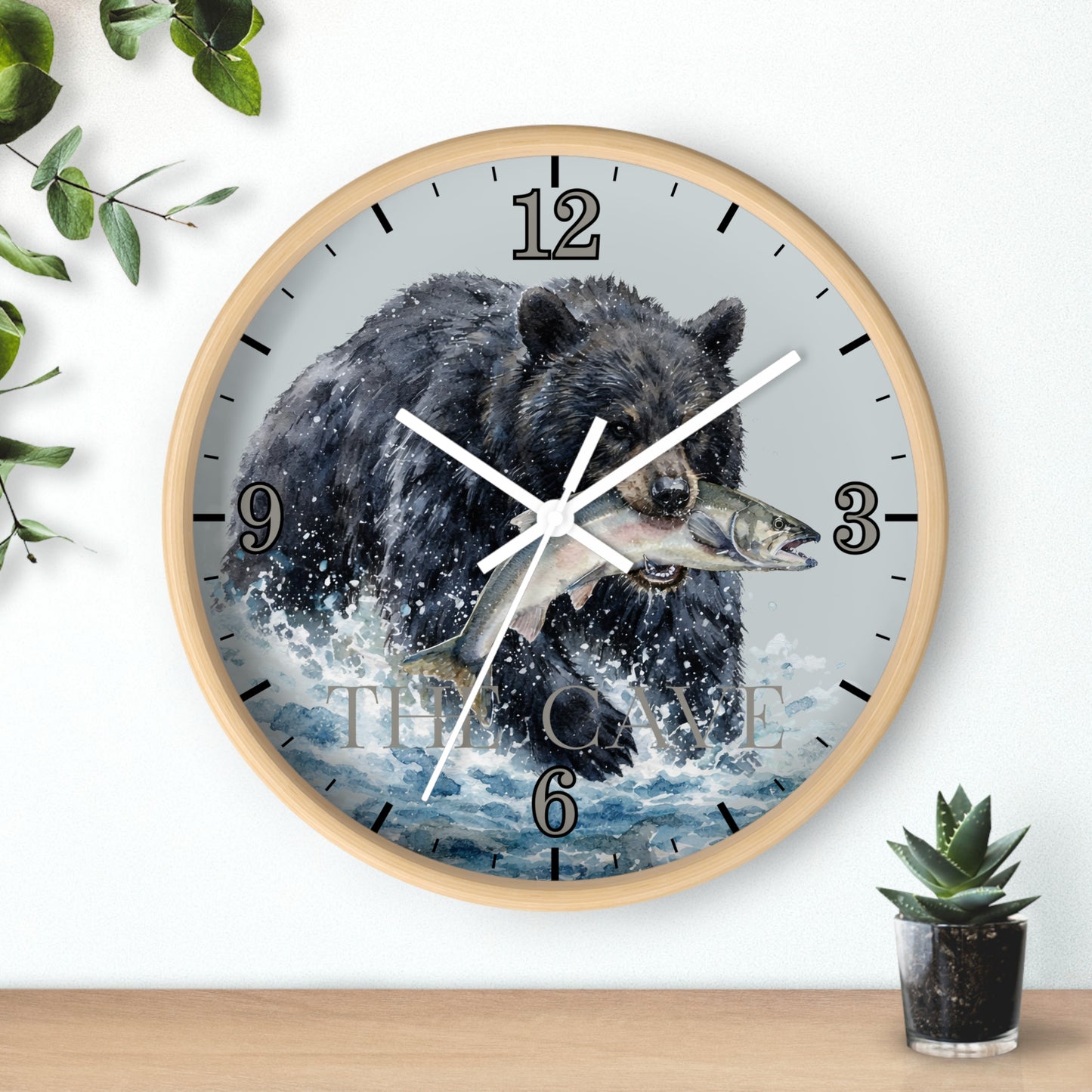 A round wall clock with a light wood frame, featuring a printed illustration of a black bear catching a fish in splashing water. The clock is mounted on a white wall above a wooden shelf, with decorative green leaves and a small potted succulent nearby.