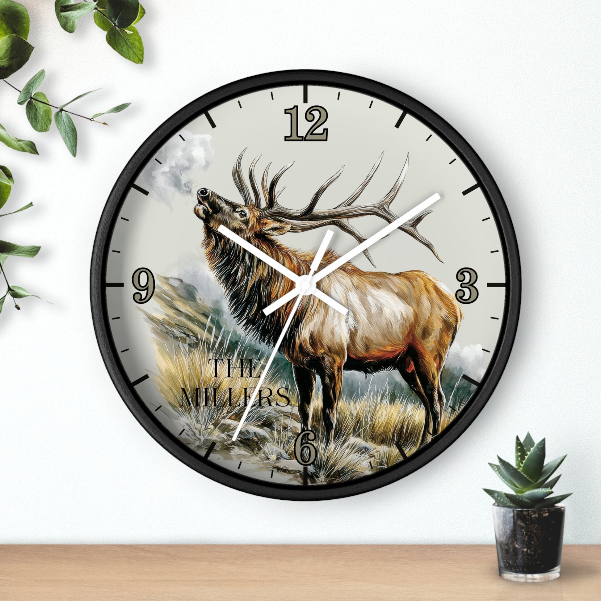 A round wall clock with a black frame and a printed design of a large elk standing in a grassy landscape. The clock face has black Arabic numerals and white hands, mounted on a white wall above a wooden surface with small green plants nearby.