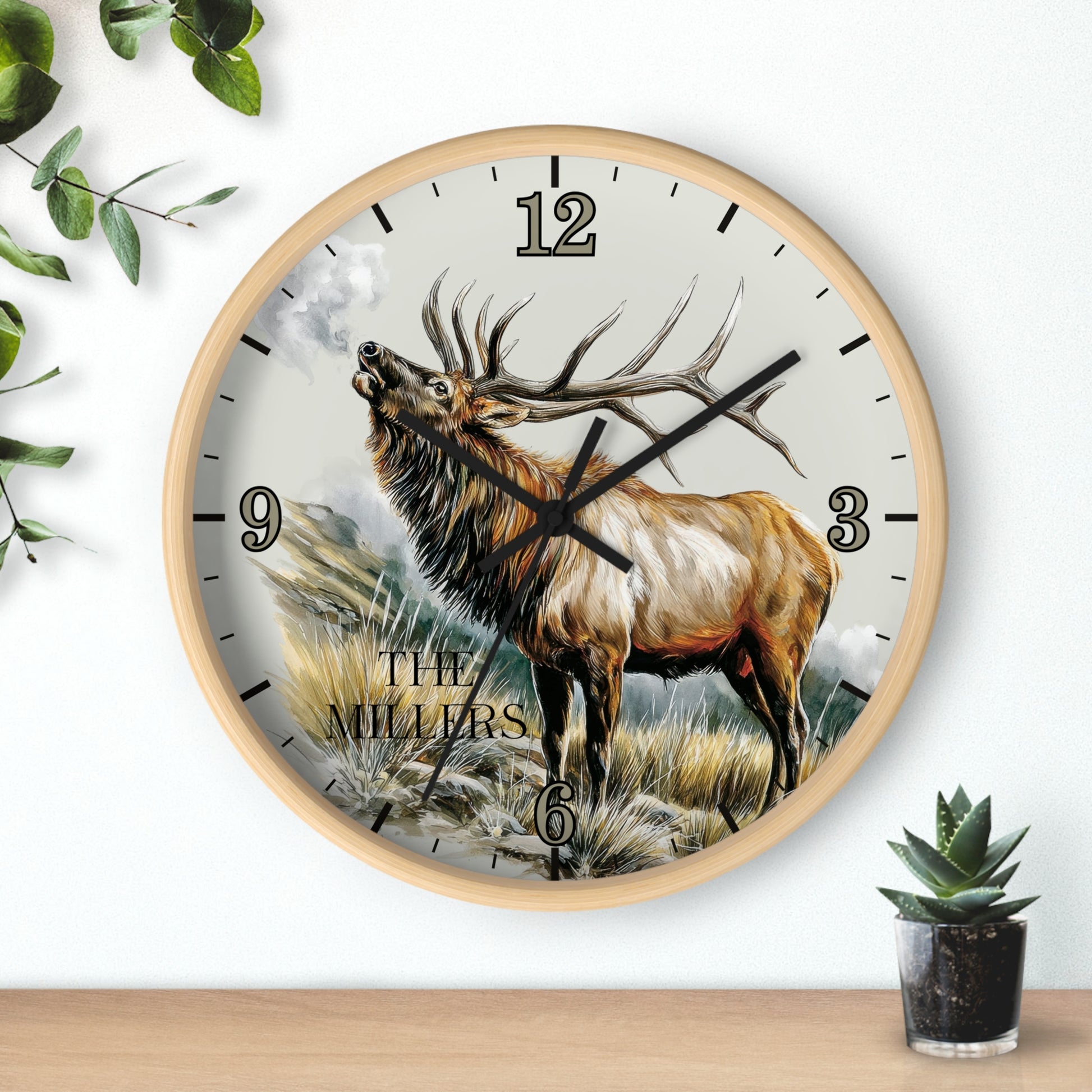A round wall clock with a light wood frame and a printed illustration of a large elk on the face. The clock is mounted on a white wall above a wooden surface, with decorative green leaves on the left and a small potted succulent on the right.
